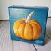 Small square painting of little orange pumpkin with teal blue background.