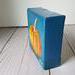 Small square painting of little orange pumpkin with teal blue background.