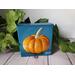 Small square painting of little orange pumpkin with teal blue background.
