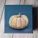 Photo of small square canvas painting of peach colored pumpkin with denim blue colored background