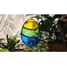 Stained glass Easter Egg suspended over a potted plant ,  additional plants in the background, Egg features yellow, green, and blue stripes