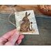 A handmade gift tag. It is a piece of real birch bark with a woodland rabbit wood burned onto the bark. A jute cord is tied in the corner. 
