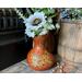 This shows a carved gourd vase with faux flowers. The vase is decorating a fireplace hearth. It sits next to an old wood barrel. 