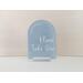 Favors please take one acrylic arch sign on clear acrylic stand on a table, with light blue paint and with white lettering.