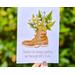 Hand holding a greeting card in the air. The card has a watercolor graphic of a hiking boot filled with wildflowers.