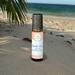 A "Beach Vibes" roll-on wellness blend by Whole Self Aromatherapy on a sandy beach with palm frond and the ocean in the blurred background.