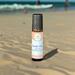 Whole Self Aromatherapy's Beach Vibes roll-on wellness blend on a sandy beach with the ocean and people swimming in the blurred background.