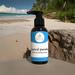 Bottle of Tropical Paradise hand and body lotion on a sandy beach with a blurry view of the ocean and palm trees in the background.