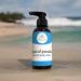 Bottle of Tropical Paradise hand and body lotion on a wet, sandy beach with a blurry view of the ocean and palm trees in the background