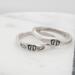 Two sterling silver rings with hand-stamped baby feet and wings, placed on a white wooden surface with delicate white flowers.
