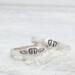 Two sterling silver rings with hand-stamped baby feet and wings, placed on a white wooden surface with delicate white flowers.
