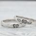 Two sterling silver rings with hand-stamped baby feet and wings, placed on a white wooden surface with delicate white flowers.
