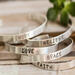 Five sterling silver bracelets with words HOPE, LOVE, GRACE, FAITH, BELIEVE, on a wood surface with a green succulent in the background.
