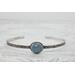 Silver bracelet with floral engravings, featuring a oval blue aquamarine on a light wood surface with scattered white baby's breath flower.

