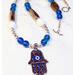 Image is of a large Hamsa hand necklace with blue evil eye in the center. The necklace has blue and white glass beads