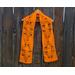 Orange scarf with leaves along the length. It is on a hanger in front of a wooden fence.