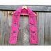 Pink Mom Crochet Scarf display on a hanger in front of a wooden fence.