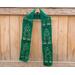 Green scarf with trees along the length. It is on a hanger in front of a wooden fence.