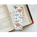 the sloth bookmark has a white background with two sloths hanging from the branches, stems of leaves, blue flowers and leaves all over the bookmark
