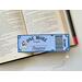 This bookmark looks like a ticket in color dark and light blue. In the center says 'One More Chapter Readers,' 'Lifetime Member' with a girl reading a book on the left and light blue flower. there are gold star boarders at each corner. Under the bar code the words 'ADMIT ONE'