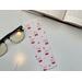 this pink bookmark is on white table with an open in the top right, and on the left is a pair of glasses and fairy lights