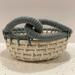A small round woven cream basket with a laurel green decorative knot design on the rim.