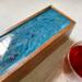 Reflective metallic ribbons of gold swirl in the shades of blue on top of this tea caddy that holds up to 50 tea bags.  This tea box is handmade of mango wood with a hinged top and has 5 compartments for organizing your tea bags.  Perfect gift for tea lover or someone who loves to entertain.  Adds a pop of color and luxury to any home decor.
