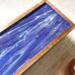 Framed by wood, the design on the top of this tea caddy is cheerful and bold.  Purple and white swirl together in ribbons on the cover of this tea box that holds up to 50 tea bags.  Tea connoisseurs will love this colorful way to display and sort their teas.