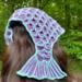 A woman with dark brown hair wearing a mermaid tail shaped crochet hair bandana that has medium purple scales with light blue outlines.