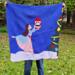 A woman holding a crochet baby blanket with a picture of a ballerina dancing with a Nutcracker doll in the snow on it. 