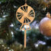 A handcrafted wooden Christmas tree ornament shaped like a lollipop, featuring intricate cut-out spiral patterns on the circular top and a simple stick base. The ornament is light brown with a smooth finish.