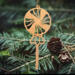 A handcrafted wooden Christmas tree ornament shaped like a lollipop, featuring intricate cut-out spiral patterns on the circular top and a simple stick base. The ornament is light brown with a smooth finish.