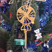 A handcrafted wooden Christmas tree ornament shaped like a lollipop, featuring intricate cut-out spiral patterns on the circular top and a simple stick base. The ornament is light brown with a smooth finish.