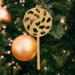 A handmade Christmas tree ornament crafted from light brown maple hardwood plywood, shaped like a lollipop with a round head featuring intricate spiral cutouts and a long stick handle, suspended from a small metal hook.