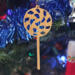 A handmade Christmas tree ornament crafted from light brown maple hardwood plywood, shaped like a lollipop with a round head featuring intricate spiral cutouts and a long stick handle, suspended from a small metal hook.