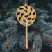 A handmade Christmas tree ornament crafted from light brown maple hardwood plywood, shaped like a lollipop with a round head featuring intricate spiral cutouts and a long stick handle, suspended from a small metal hook.