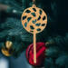 A handmade Christmas tree ornament crafted from light brown maple hardwood plywood, shaped like a lollipop with a round head featuring intricate spiral cutouts and a long stick handle, suspended from a small metal hook.