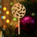 A handmade Christmas tree ornament crafted from light brown maple hardwood plywood, shaped like a lollipop with a round head featuring intricate spiral cutouts and a long stick handle, suspended from a small metal hook.