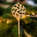 A handmade Christmas tree ornament crafted from light brown maple hardwood plywood, shaped like a lollipop with a round head featuring intricate spiral cutouts and a long stick handle, suspended from a small metal hook.