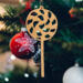 A handmade Christmas tree ornament crafted from light brown maple hardwood plywood, shaped like a lollipop with a round head featuring intricate spiral cutouts and a long stick handle, suspended from a small metal hook.