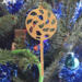 A handmade Christmas tree ornament crafted from light brown maple hardwood plywood, shaped like a lollipop with a round head featuring intricate spiral cutouts and a long stick handle, suspended from a small metal hook.