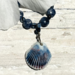 Closeup of ceramic shell pendant with blue and brown ridges attached to a string of blue sodalite beads peeking out from under white petals.