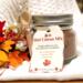 Handmade hot cocoa mix jar with wooden spoon and red-gold ribbon, displayed on a white knit blanket with autumn leaves.