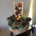 Rustic holiday centerpiece with pinecones, greenery, and a glass vase filled with gold and bronze ornaments on a wood-slice base.