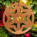 A handmade wooden Christmas tree ornament, circular in shape, crafted from light brown wood with a natural grain pattern. It features an intricate, symmetrical cutout design resembling a star or snowflake, with multiple pointed shapes radiating from a central hole. The ornament is attached to a small metal hook for hanging, showcasing its rustic and festive craftsmanship.