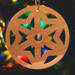 A handmade wooden Christmas tree ornament, circular in shape, crafted from light brown wood with a natural grain pattern. It features an intricate, symmetrical cutout design resembling a star or snowflake, with multiple pointed shapes radiating from a central hole. The ornament is attached to a small metal hook for hanging, showcasing its rustic and festive craftsmanship.