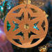 A handmade wooden Christmas tree ornament, circular in shape, crafted from light brown wood with a natural grain pattern. It features an intricate, symmetrical cutout design resembling a star or snowflake, with multiple pointed shapes radiating from a central hole. The ornament is attached to a small metal hook for hanging, showcasing its rustic and festive craftsmanship.
