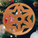 A handmade wooden Christmas tree ornament, circular in shape, crafted from light brown wood with a natural grain pattern. It features an intricate, symmetrical cutout design resembling a star or snowflake, with multiple pointed shapes radiating from a central hole. The ornament is attached to a small metal hook for hanging, showcasing its rustic and festive craftsmanship.