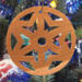 A handmade wooden Christmas tree ornament, circular in shape, crafted from light brown wood with a natural grain pattern. It features an intricate, symmetrical cutout design resembling a star or snowflake, with multiple pointed shapes radiating from a central hole. The ornament is attached to a small metal hook for hanging, showcasing its rustic and festive craftsmanship.