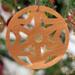 A handmade wooden Christmas tree ornament, circular in shape, crafted from light brown wood with a natural grain pattern. It features an intricate, symmetrical cutout design resembling a star or snowflake, with multiple pointed shapes radiating from a central hole. The ornament is attached to a small metal hook for hanging, showcasing its rustic and festive craftsmanship.
